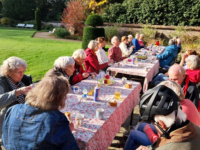 Vrijwilligers zitten aan een lange tafel in de Dieptetuin in Zeist te genieten van koffie met appeltaart